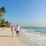 Senior couple walking barefoot on sunny beach under palm trees.